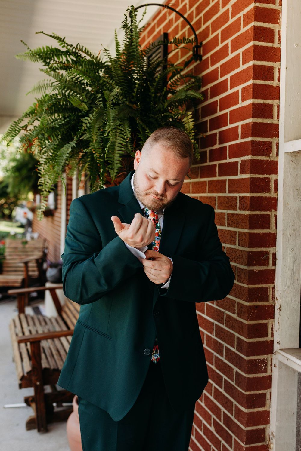 A groom in a dark green suit adjusts his cufflinks on a brick porch, wearing a colorful floral tie.