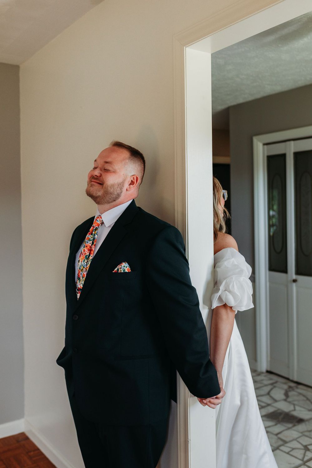 A groom in a black suit with floral tie holds hands with a bride in a white dress around a doorframe.