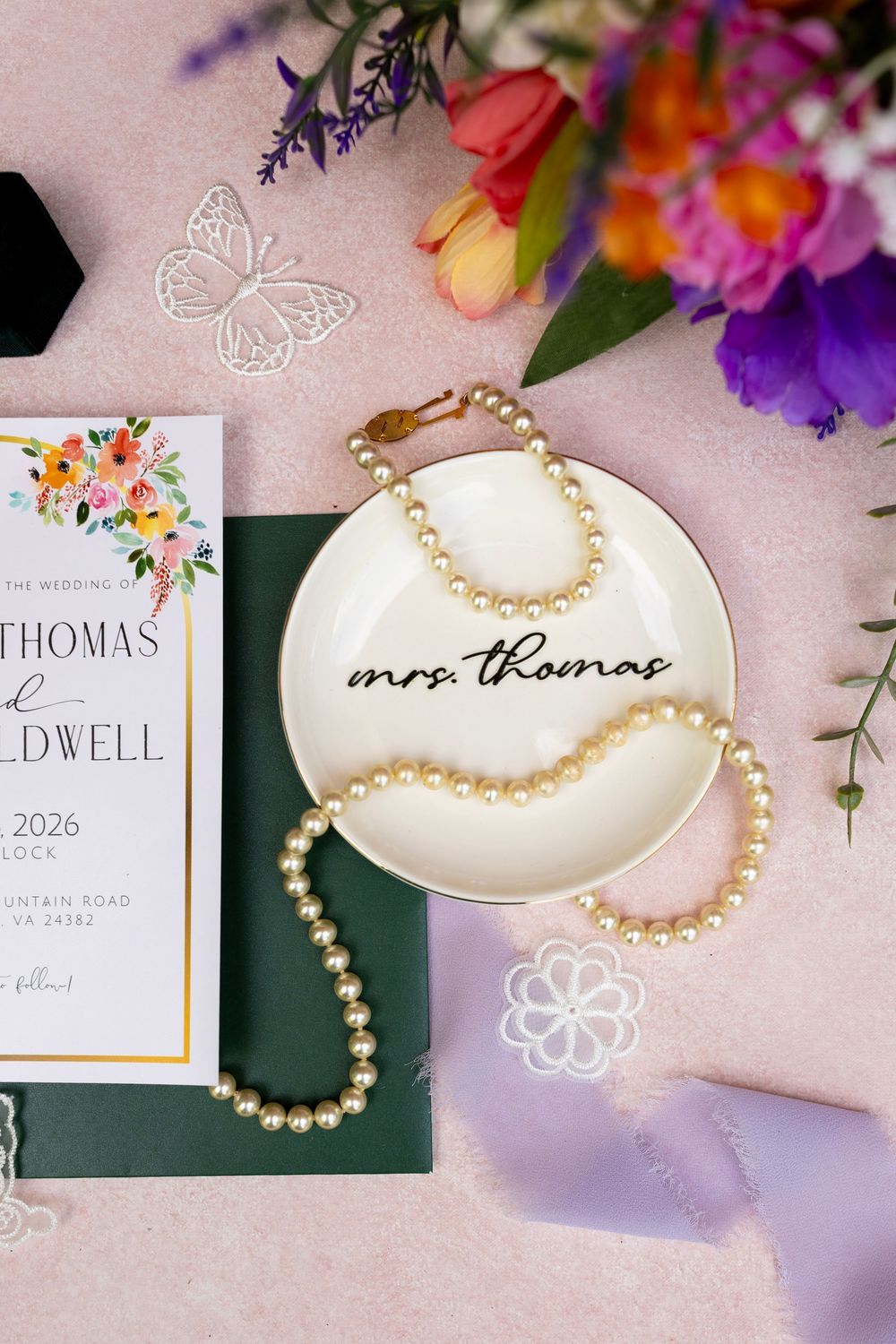 Wedding flat lay featuring pearl necklace on mrs. thomas ring dish, floral invitation suite, and colorful flowers.