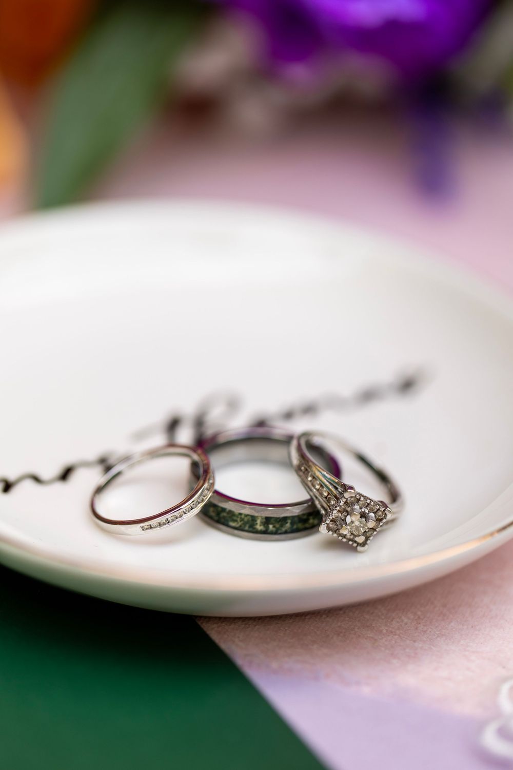 Three wedding rings displayed on a white ceramic dish, including diamond halo engagement ring and green gemstone band.