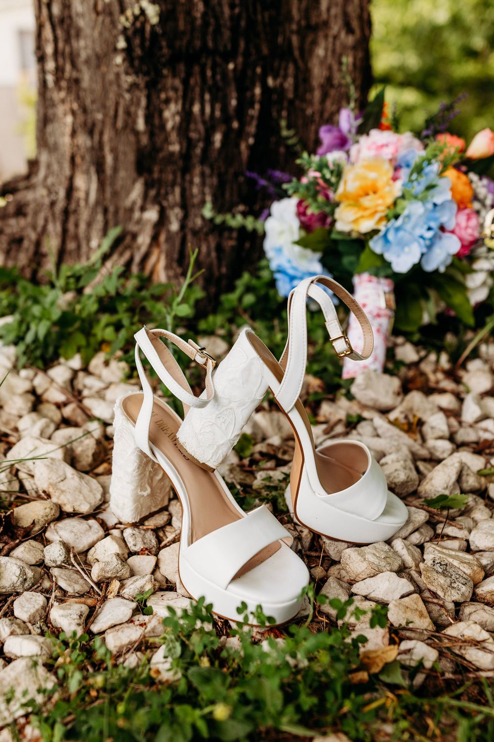 White bridal heels with lace detail on rocky ground beside colorful wedding bouquet near tree.