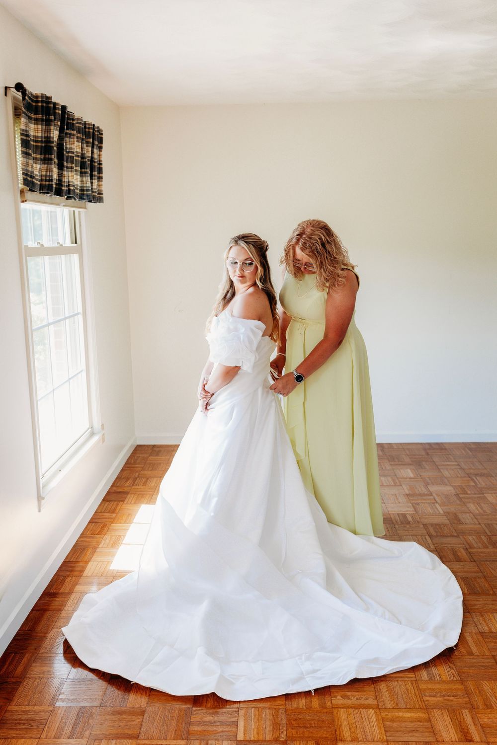 Bride in off-shoulder white gown with train, assisted by bridesmaid in sage green dress in sunlit room.