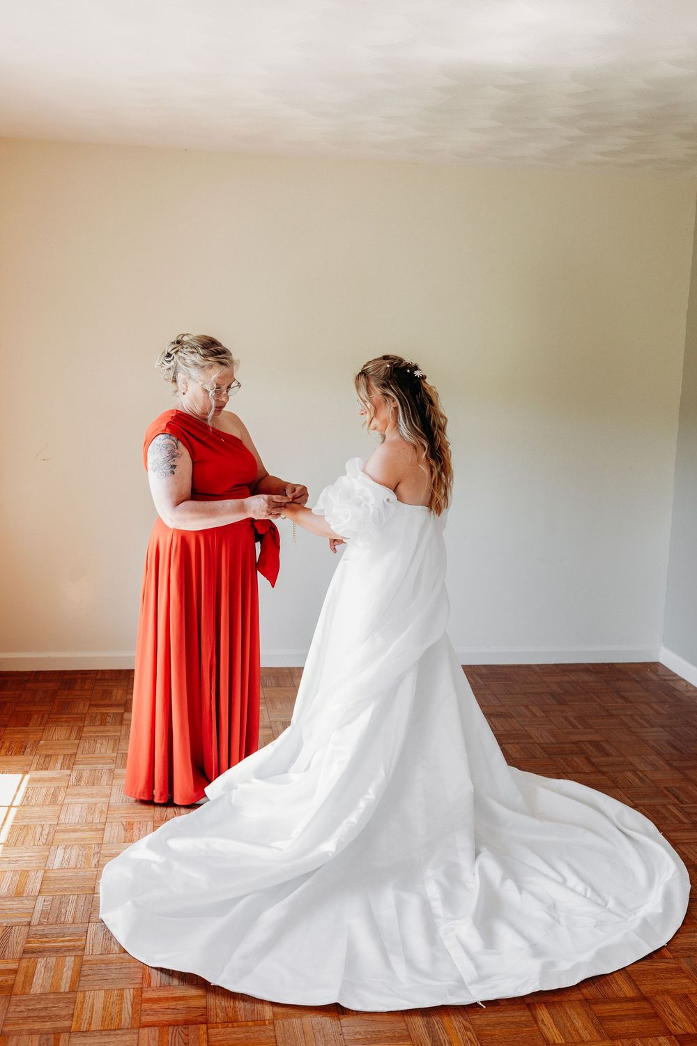 Bride in white gown being helped by bridesmaid in red dress during wedding preparation indoors.