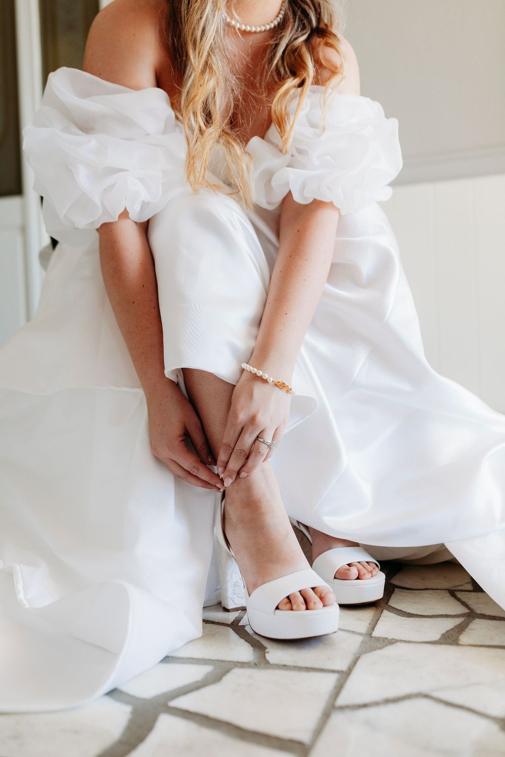 Bride in off-shoulder white gown adjusting white platform heels with pearl bracelet and ring.