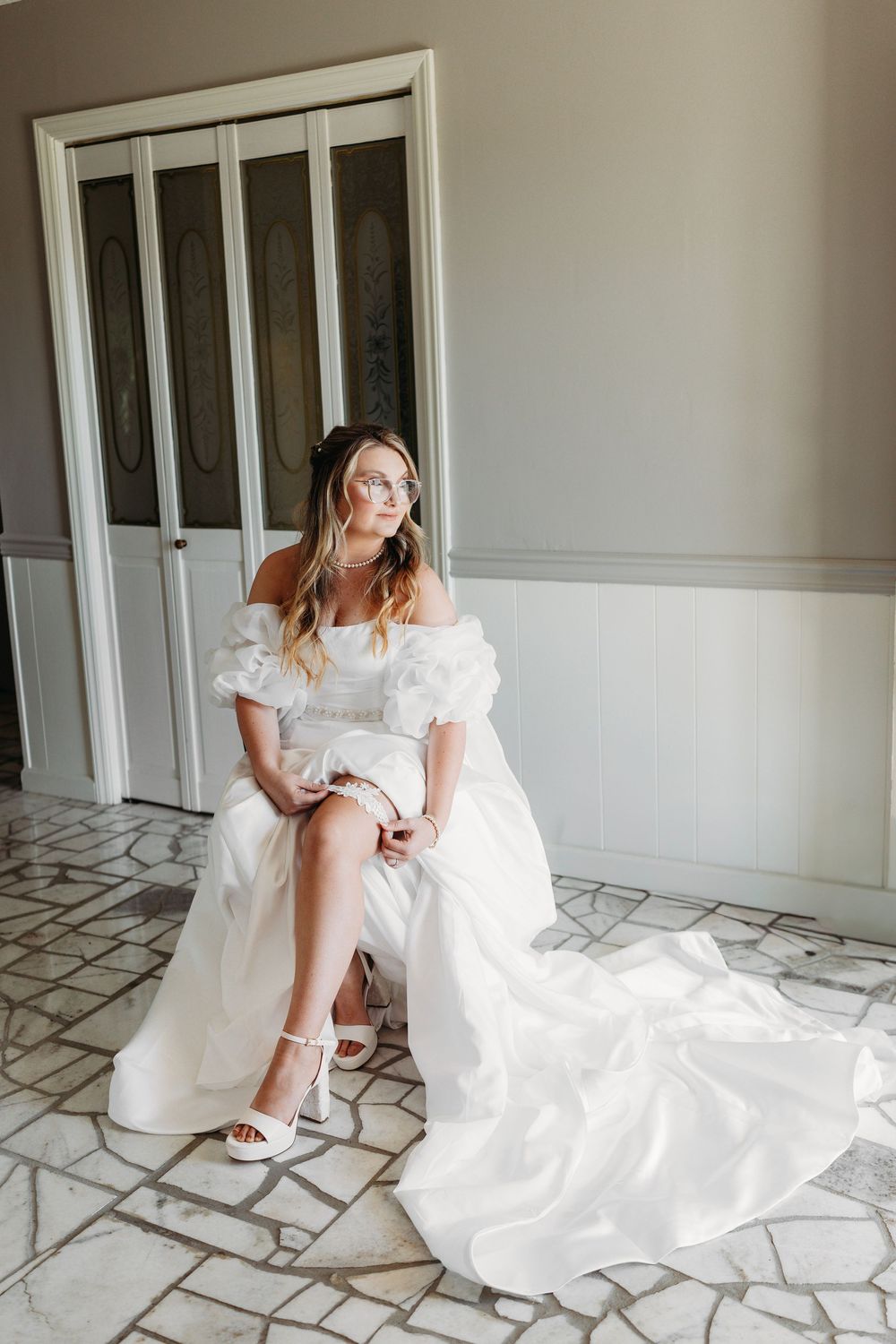 Elegant bride in off-shoulder white gown with flowing train sits gracefully on marble floor before wedding ceremony.