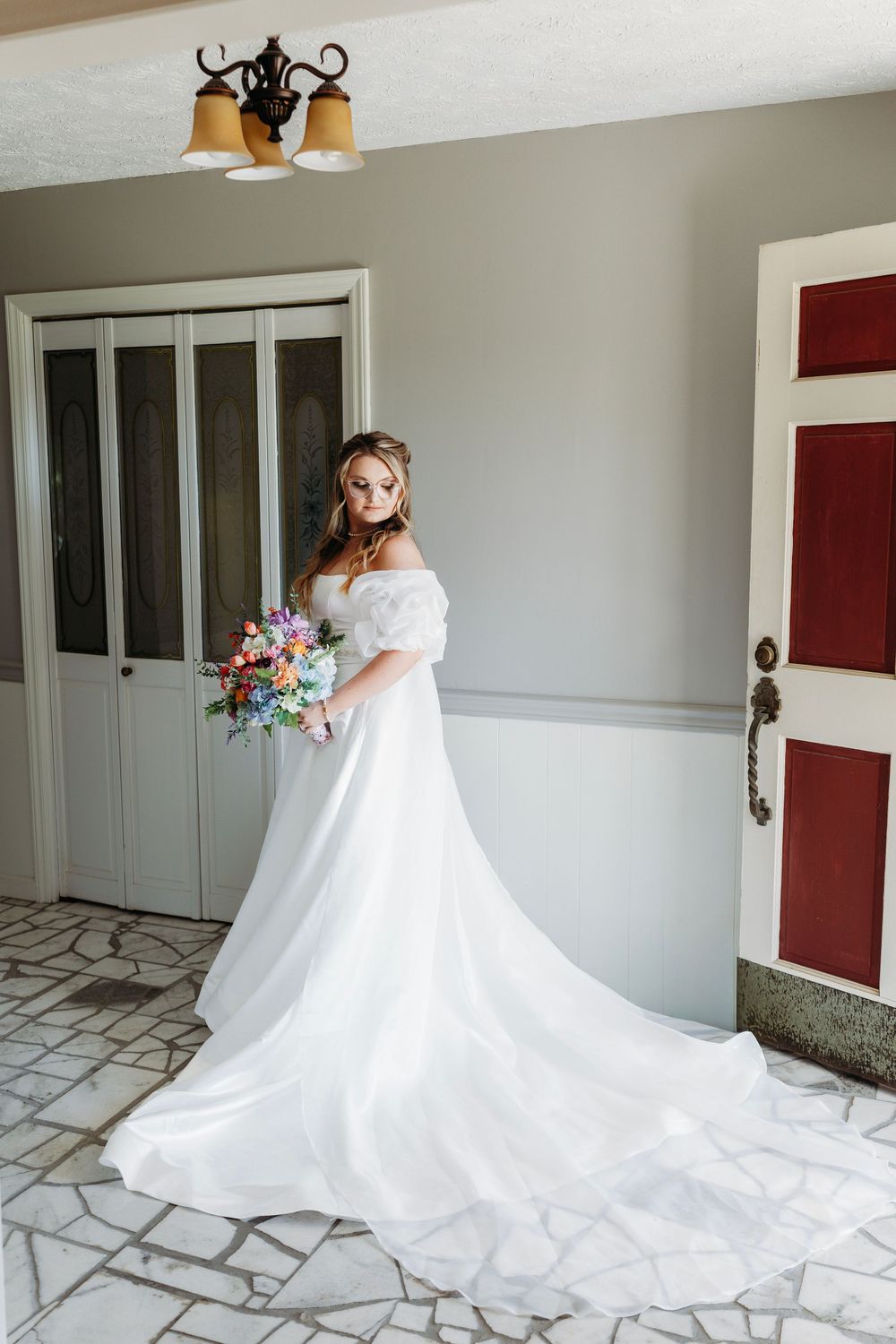 Bride in elegant off-shoulder white gown with long train holding colorful bouquet indoors.