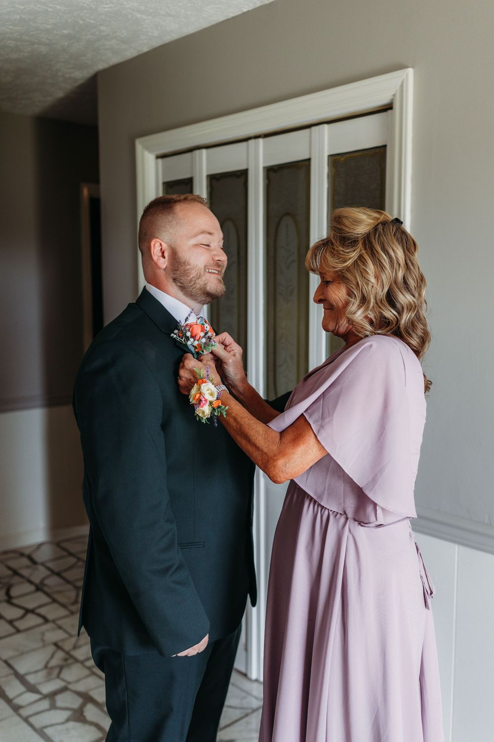 A woman in lavender dress pins a boutonniere on a smiling groom in a black suit before his wedding ceremony.