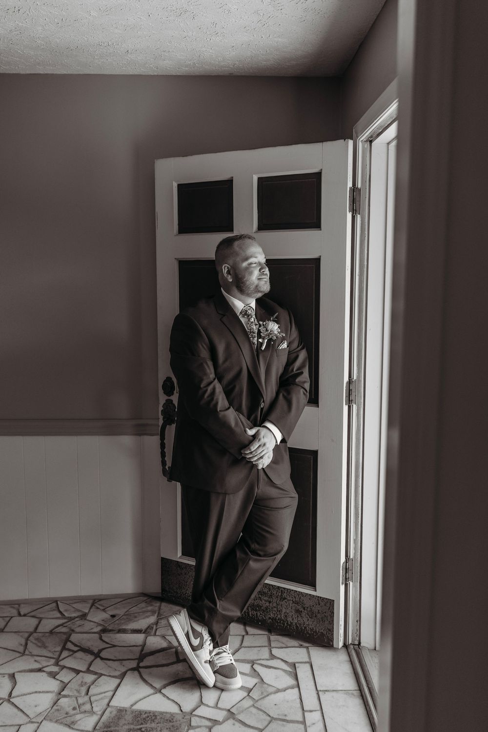 Groom in dark suit and sneakers leaning against door in black and white wedding photo.