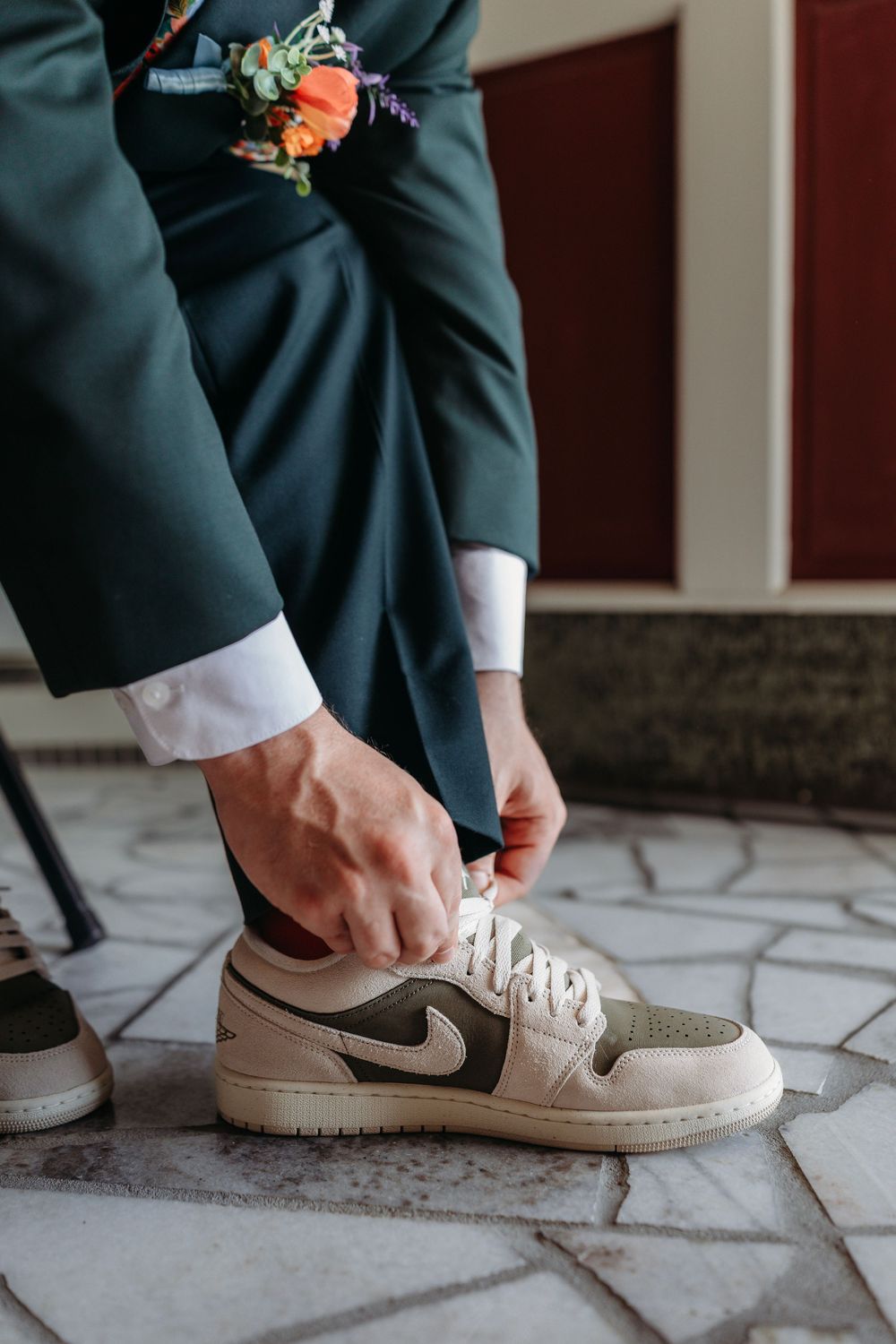 Groom in dark suit tying laces on beige Nike Air Jordan 1 sneakers with floral boutonniere visible.