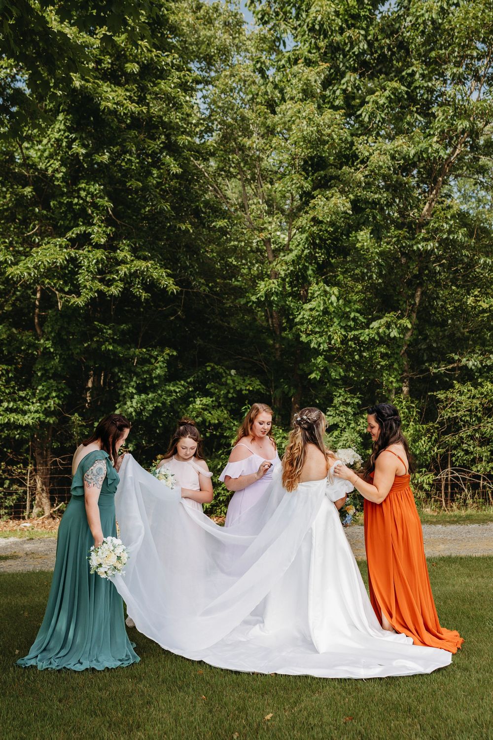 Bride in white gown with bridesmaids in teal and orange dresses arranging her train outdoors.