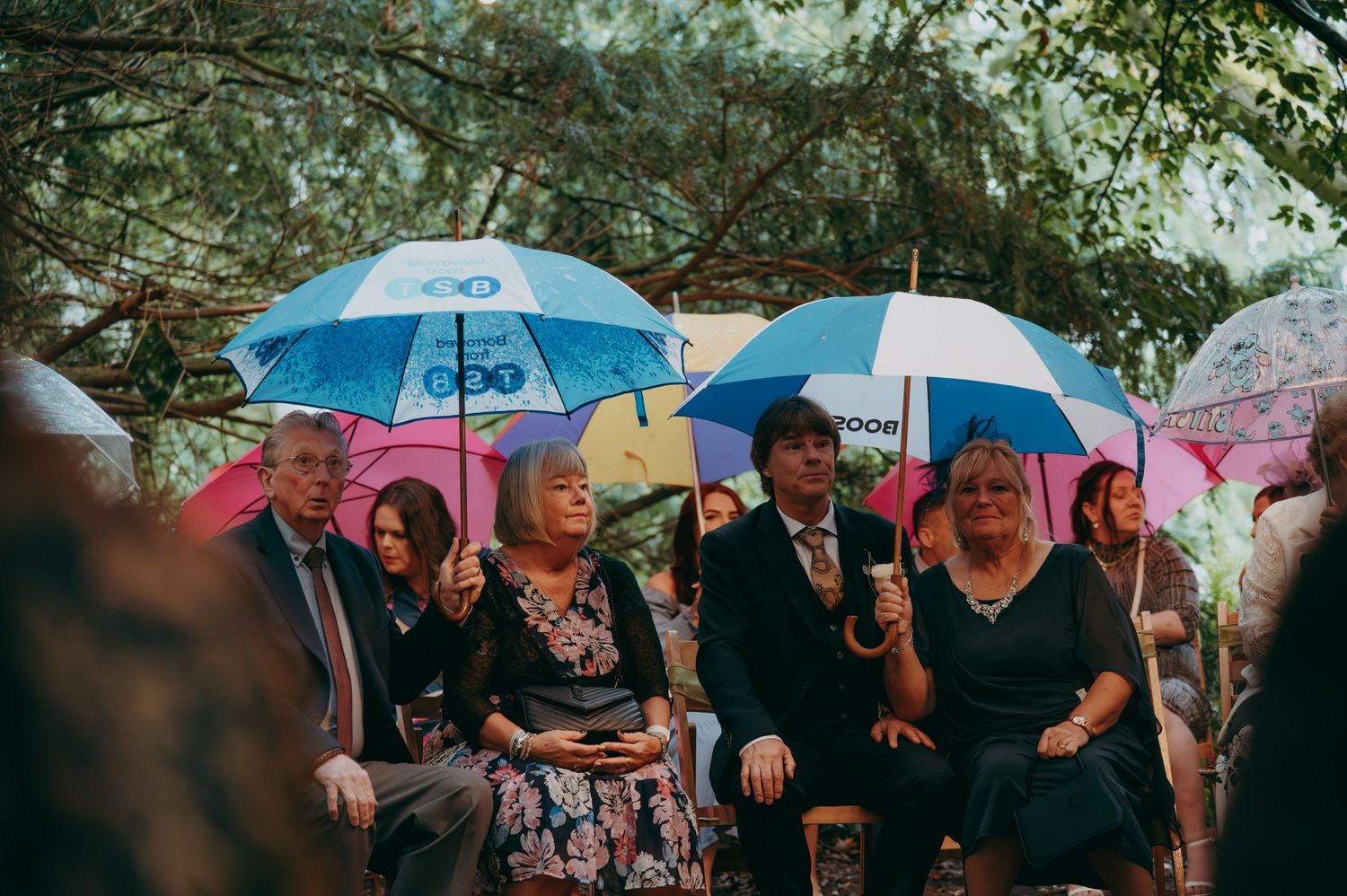 Guests seated outdoors under colorful umbrellas during a rainy wedding ceremony in a garden setting.