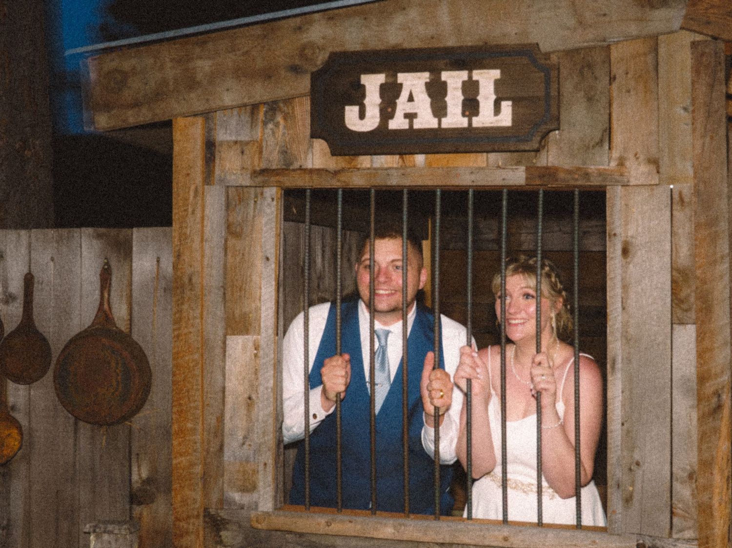 Bride and groom posing playfully behind bars of a rustic wooden jail prop at wedding.