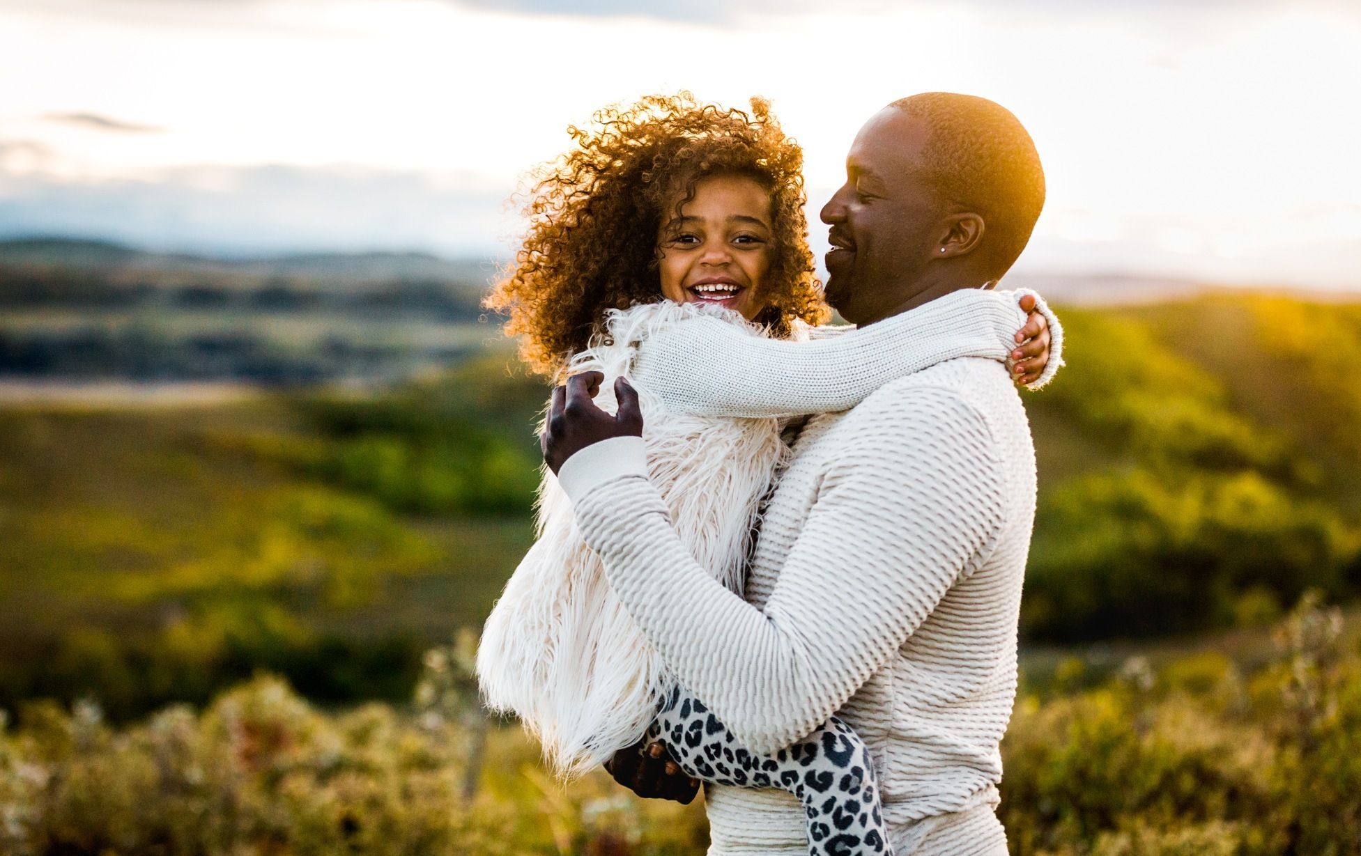 A joyful father holds his laughing curly-haired daughter outdoors with lush green hills in the background.