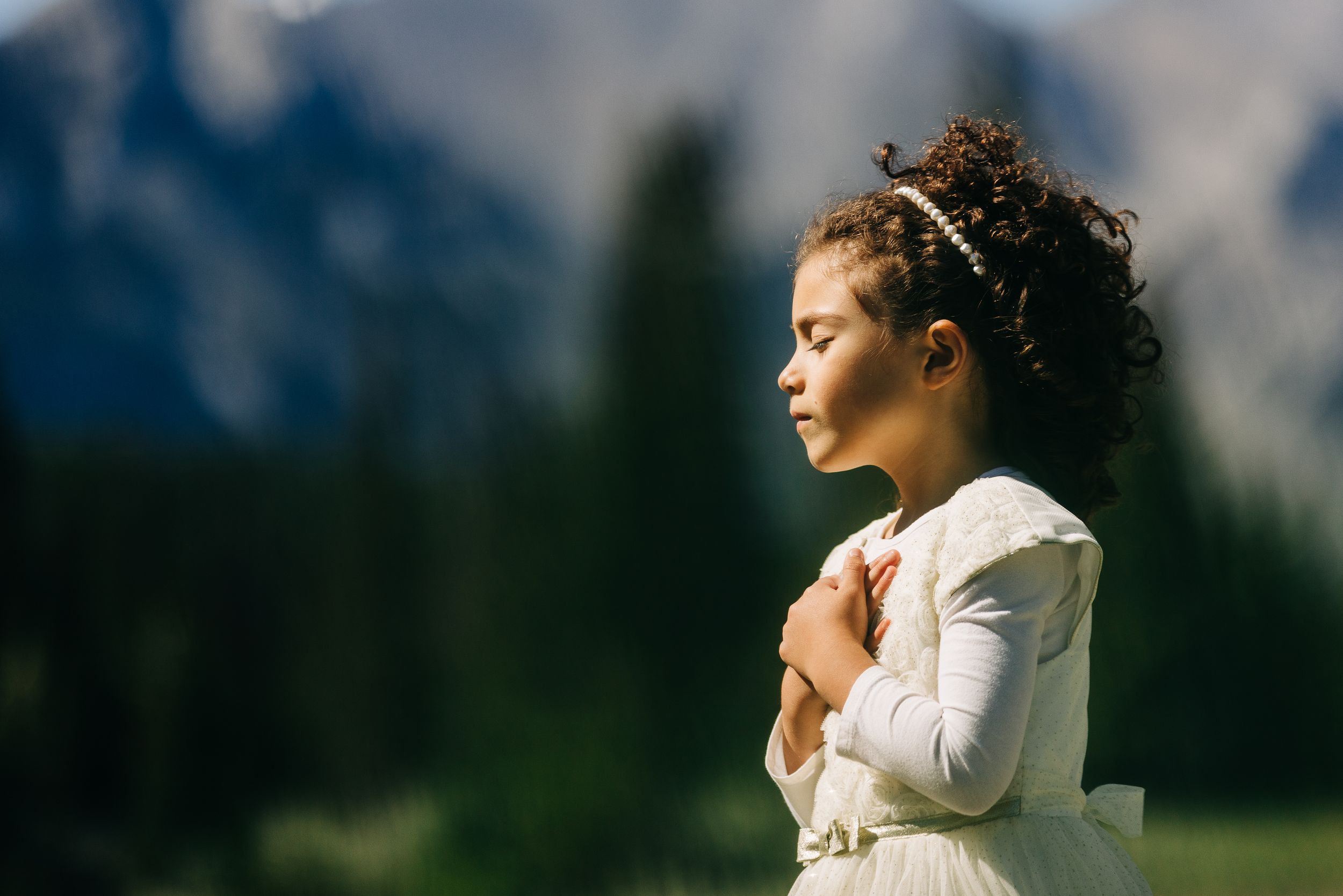 Young girl in white dress praying outdoors with mountain and forest backdrop in natural light.