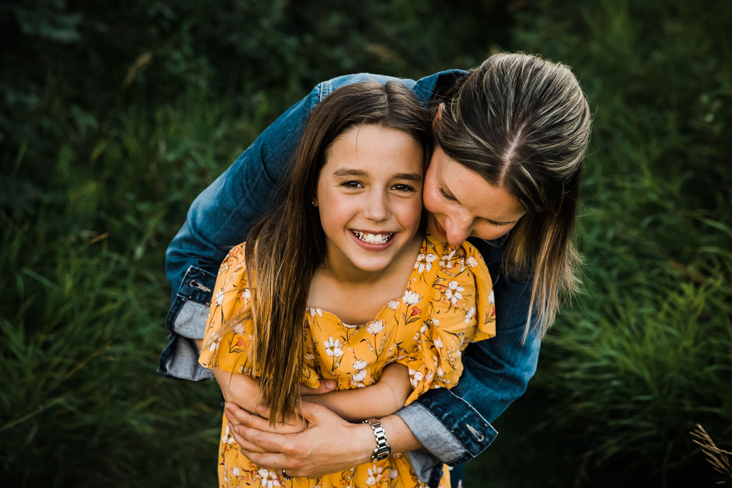 A mother hugs her smiling daughter from behind outdoors, both wearing casual clothes with green foliage in the background.