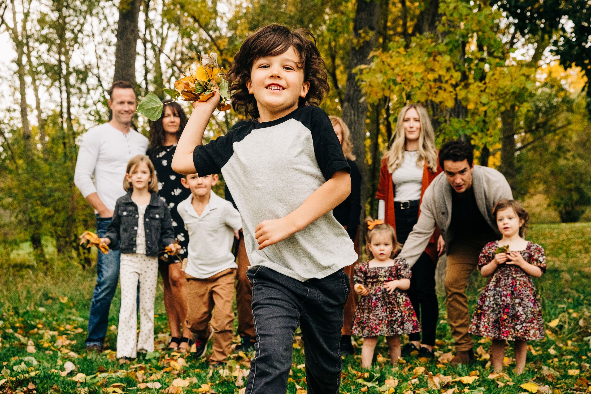 Family gathering outdoors in autumn park with children playing in fallen leaves among tall trees with golden foliage.