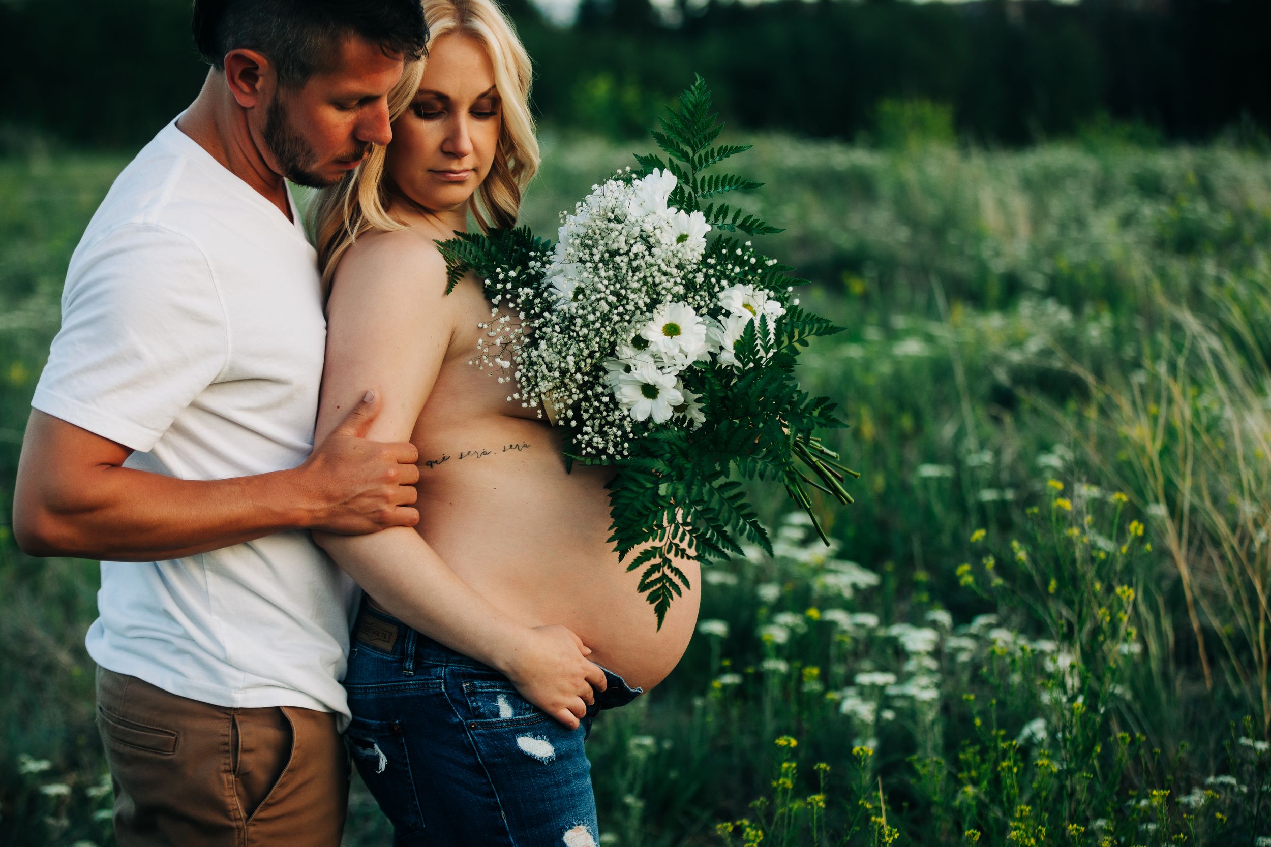 Couple in a field, pregnant woman holding white flowers bouquet, man embracing her from behind outdoors.
