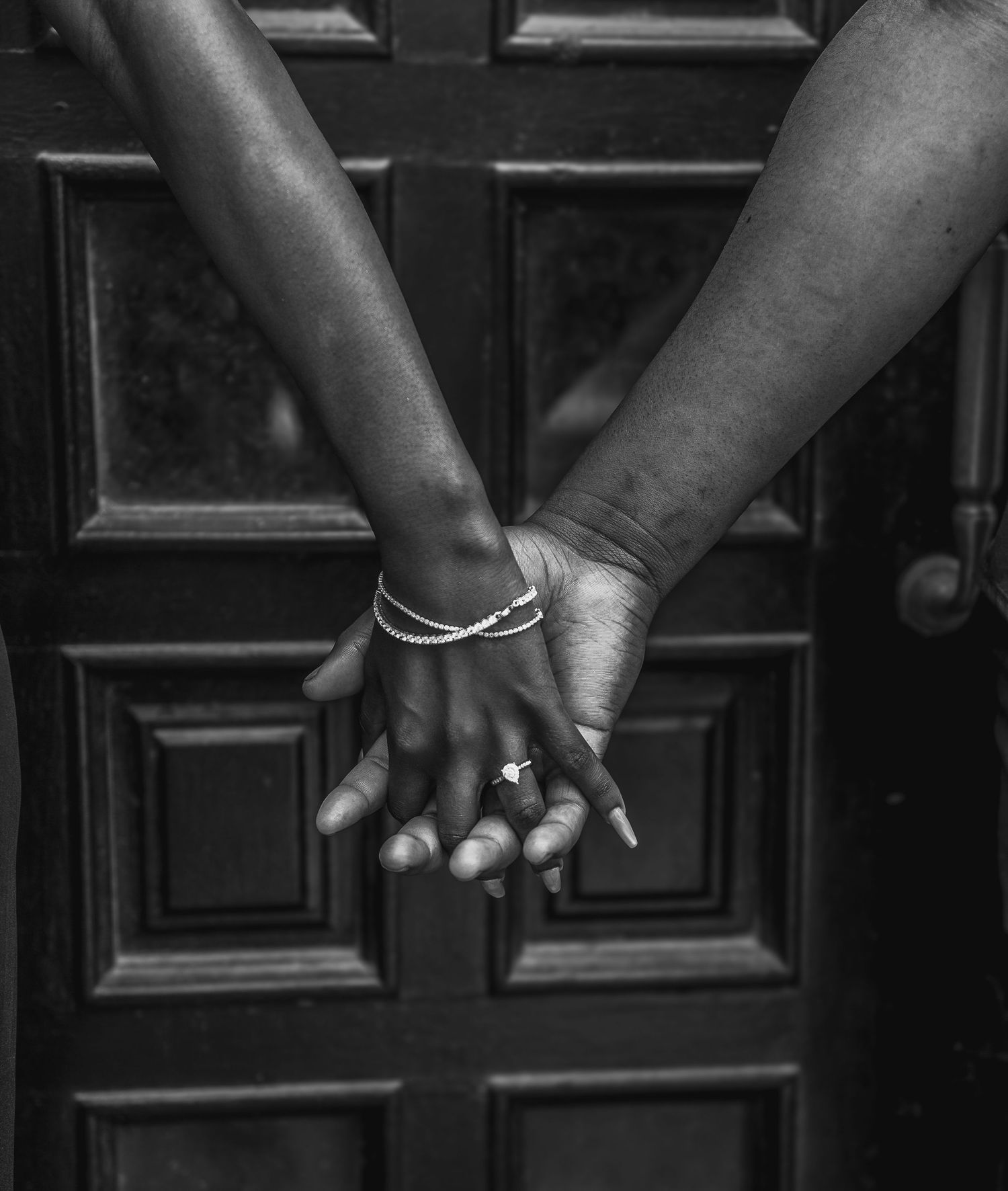 Black and white photo of two people holding hands, one wearing a diamond ring and bracelet, in front of a wooden door.