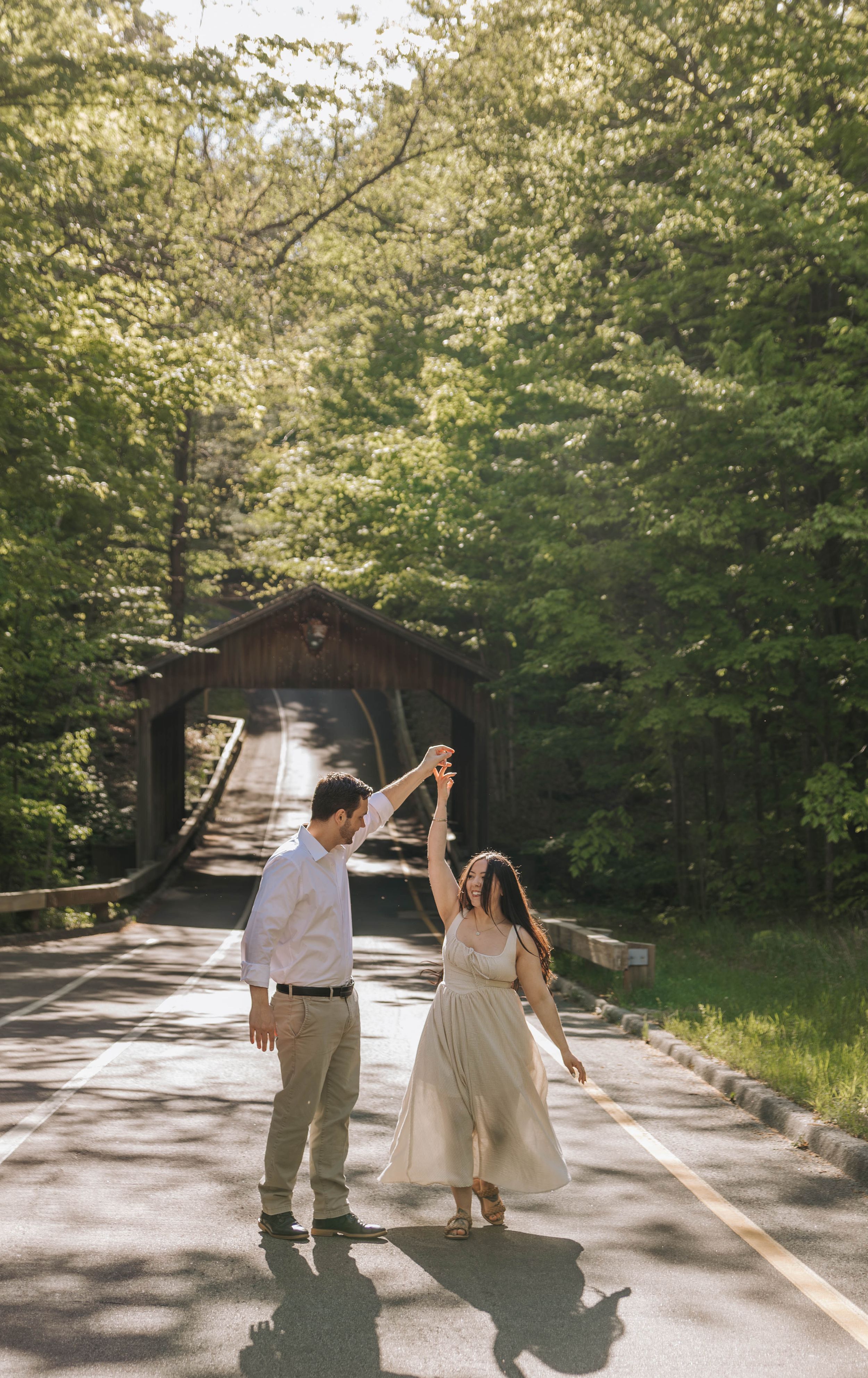 Couple dancing on a sunlit road near a rustic covered bridge surrounded by lush green trees.