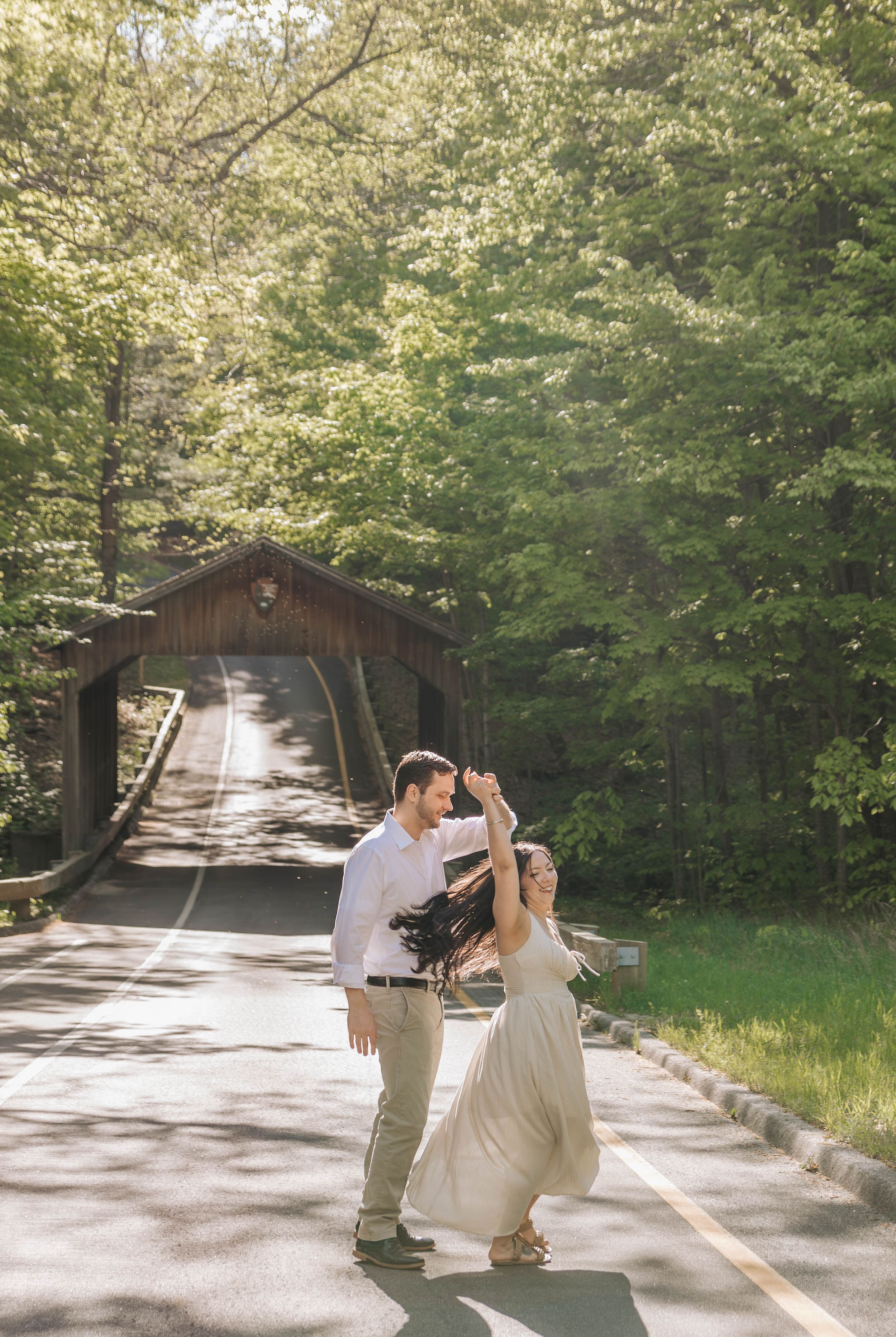 Couple dancing on a sunlit road in front of a rustic covered bridge surrounded by lush green trees.