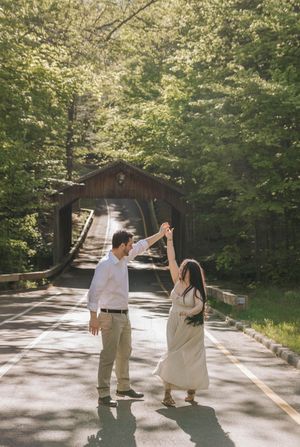 Couple dancing on a sunlit road near a rustic covered bridge surrounded by lush green trees.