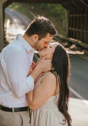 A couple shares a romantic kiss near a covered bridge, the man in a white shirt and woman in a beige dress.