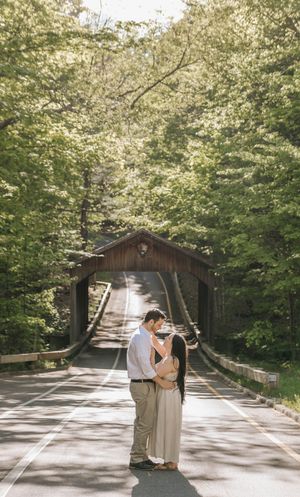 Couple sharing a romantic moment on a road leading to a rustic covered bridge surrounded by lush green trees.