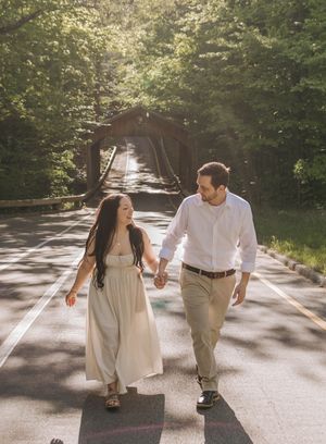 Couple holding hands walking on a sunlit road with a wooden covered bridge and lush green trees in the background.