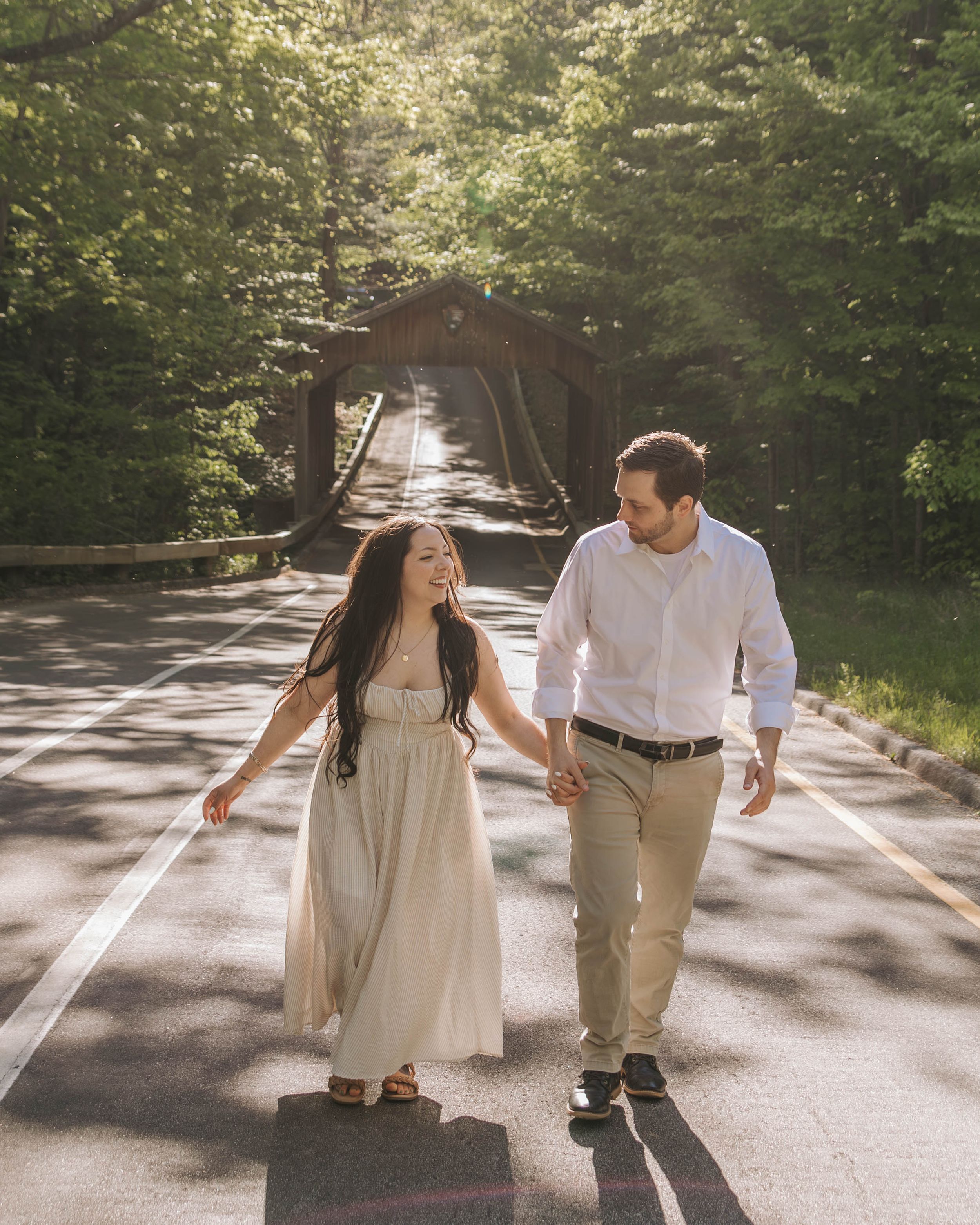 Couple holding hands walking on a road with a covered bridge and lush green trees in the background.