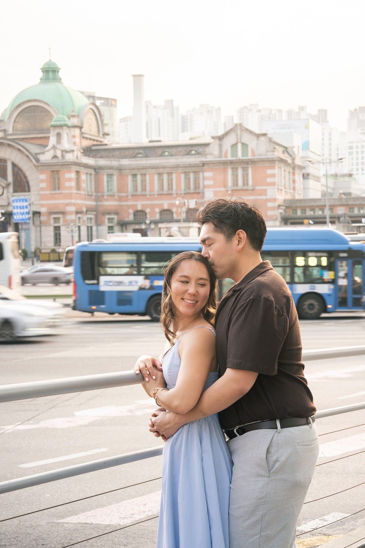 Couple embracing and smiling on a busy city street with a historic red brick building and blue bus in the background.
