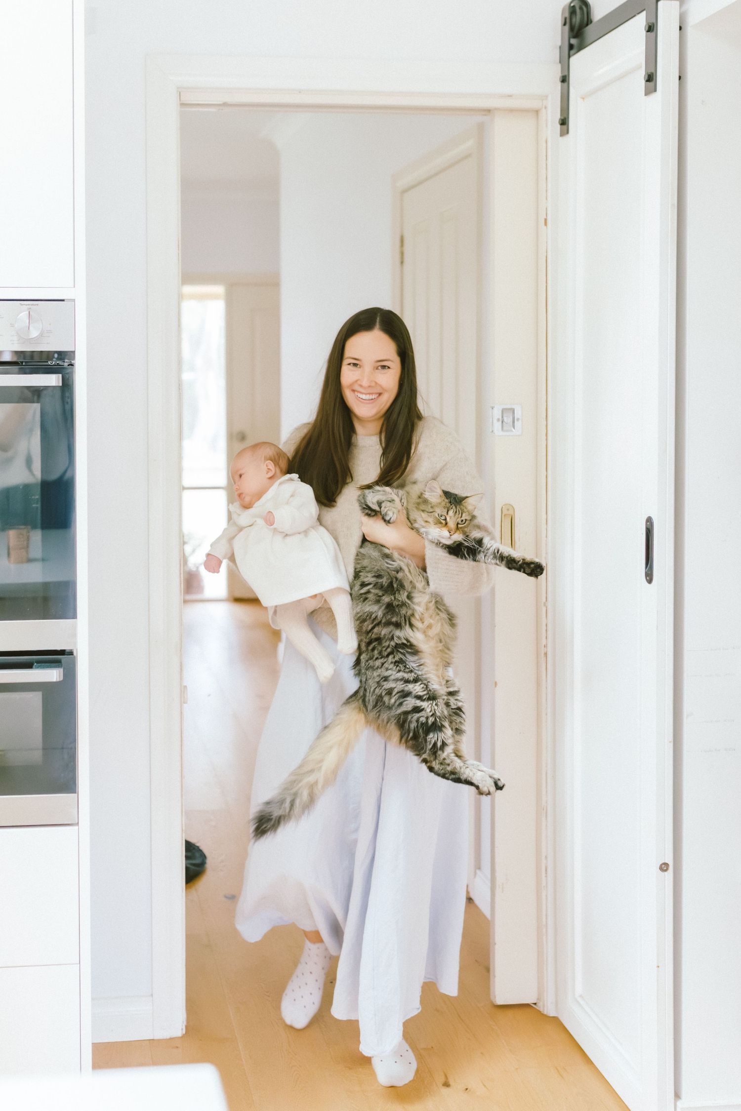 Smiling woman holding a baby and a large tabby cat while standing in a bright white kitchen doorway.