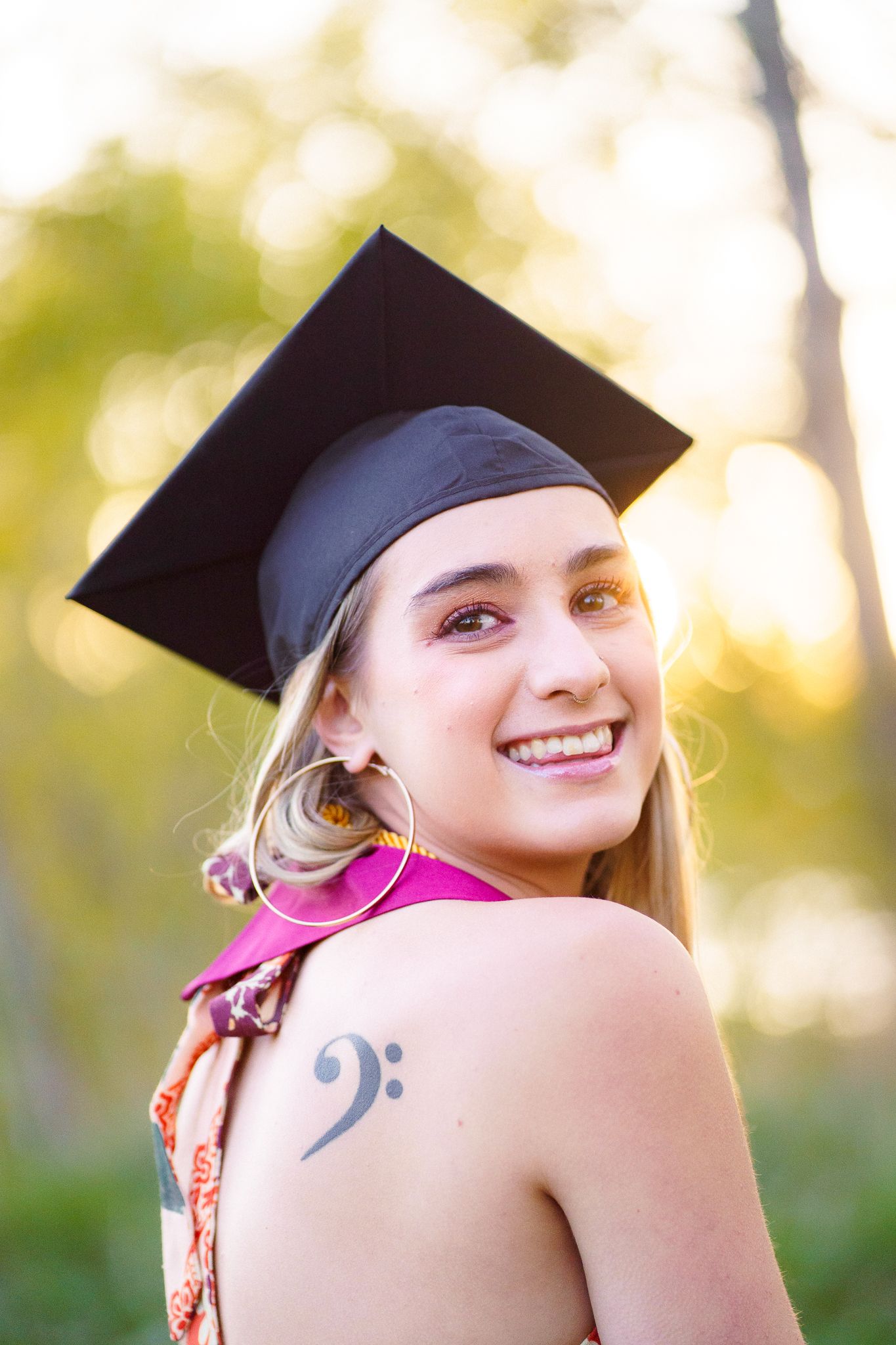 Smiling graduate wearing black cap and halter dress, showing bass clef tattoo, outdoors with golden bokeh background.