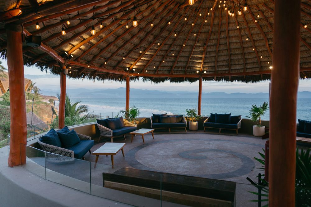 Luxury palapa lounge with blue cushioned sofas, string lights, and panoramic ocean views at a tropical resort.