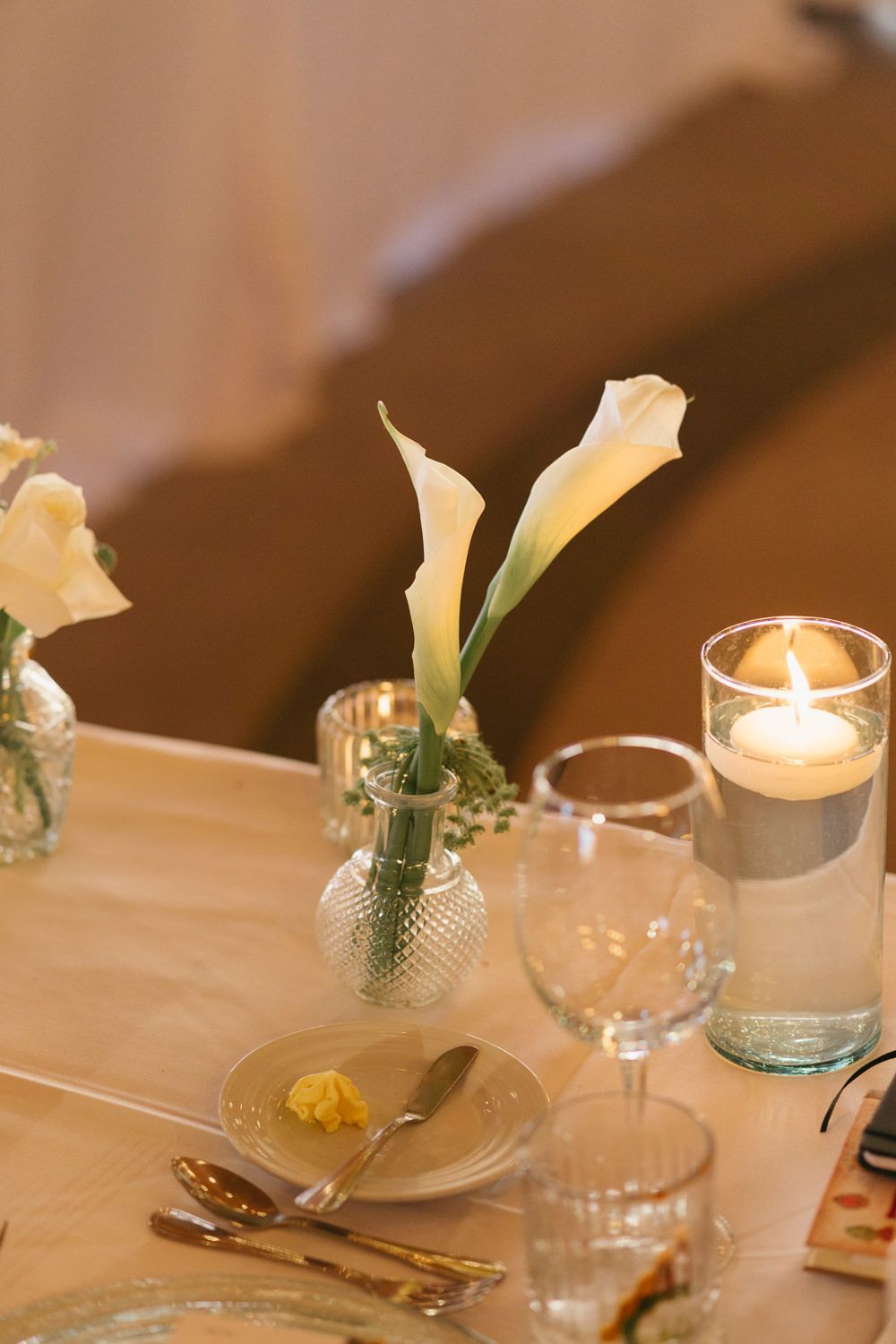 Elegant wedding table setting with white calla lilies in glass vase, lit candle, crystal glasses, and gold cutlery.