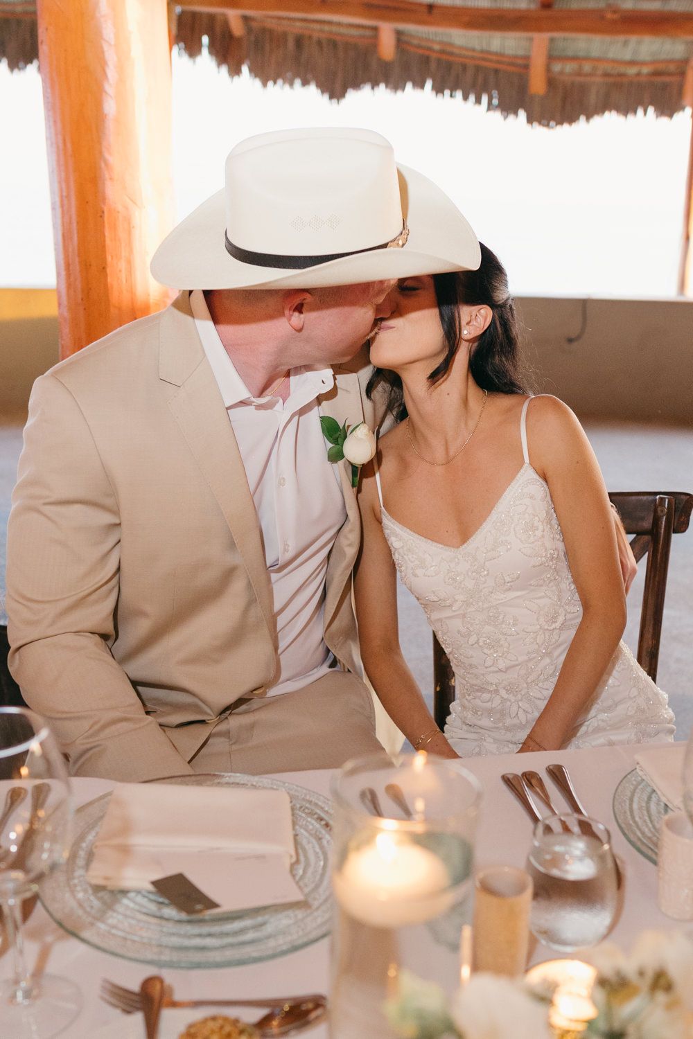 Bride and groom sharing a kiss at their wedding reception table, groom wearing a white cowboy hat and tan suit.