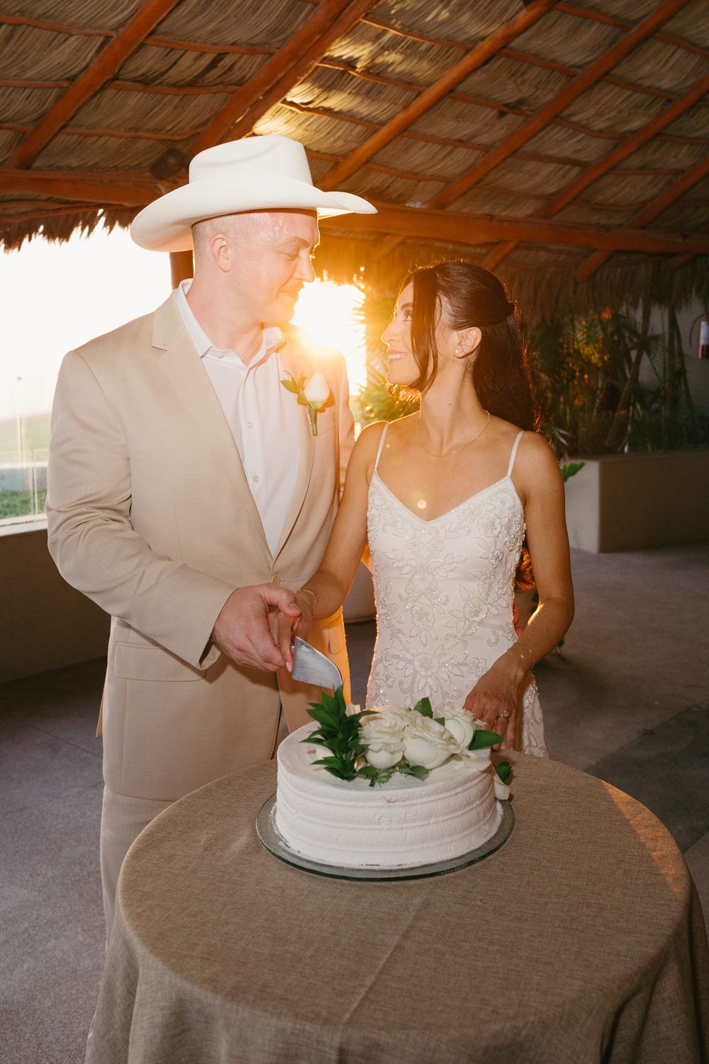 Bride and groom cutting white wedding cake at golden hour sunset under rustic thatched roof venue.