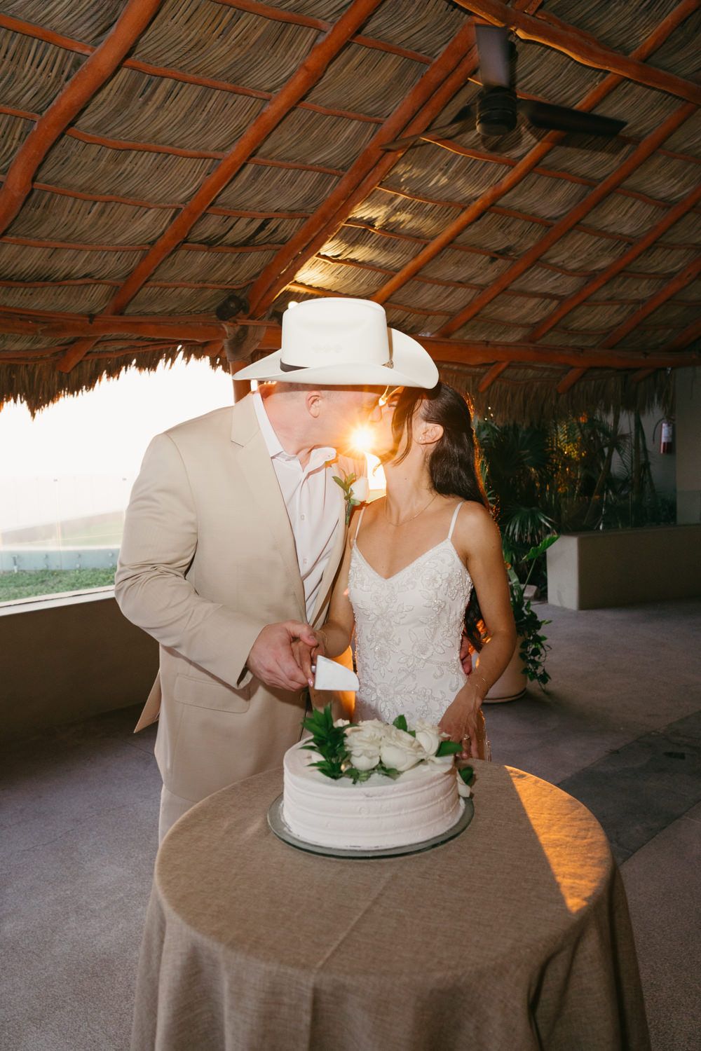 Bride and groom kissing while cutting white wedding cake under thatched palapa roof at sunset beach venue.