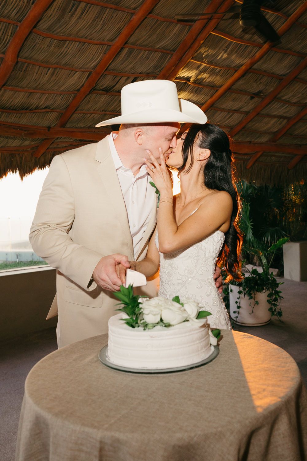 Bride and groom kissing while cutting white wedding cake under thatched roof outdoor venue.