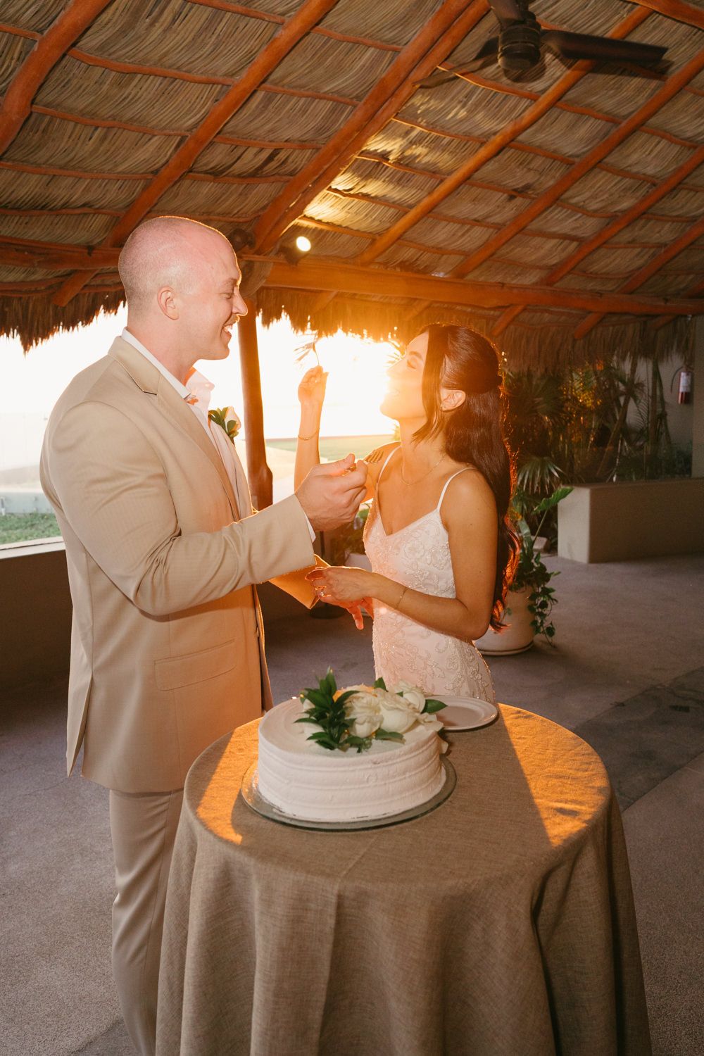 Bride and groom cutting white wedding cake at golden hour under a thatched palapa roof at a beach venue.