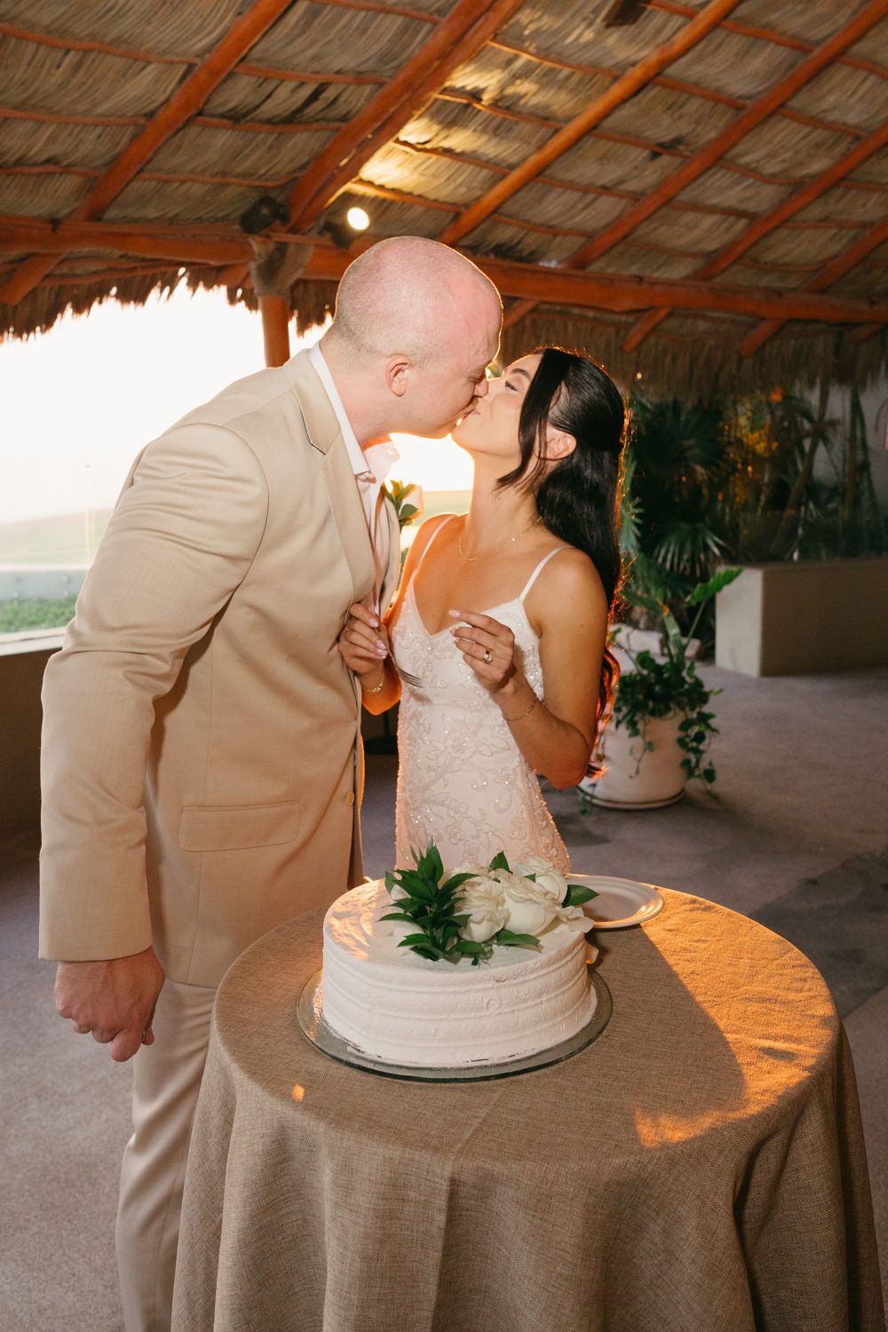 Bride and groom sharing a kiss over their white wedding cake under a tropical palapa venue at sunset.