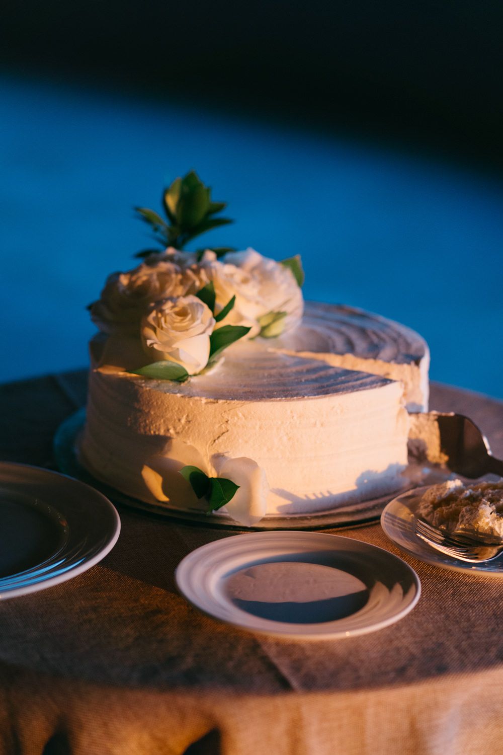 Elegant white wedding cake decorated with cream roses and green leaves on a draped table in blue evening light.