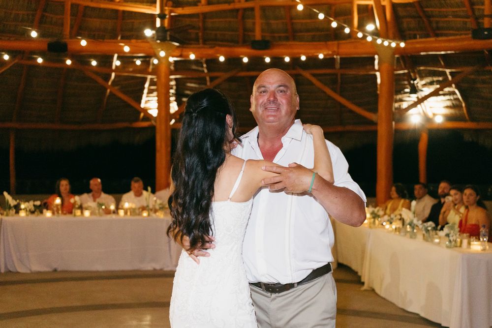 Bride and father share an emotional first dance at an outdoor wedding reception with string lights and guests seated at tables.