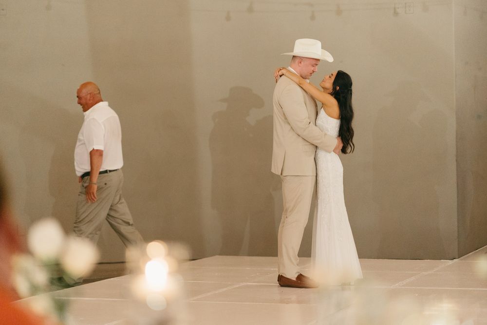 Couple shares first dance at wedding reception, groom in tan suit and white hat, bride in white gown.