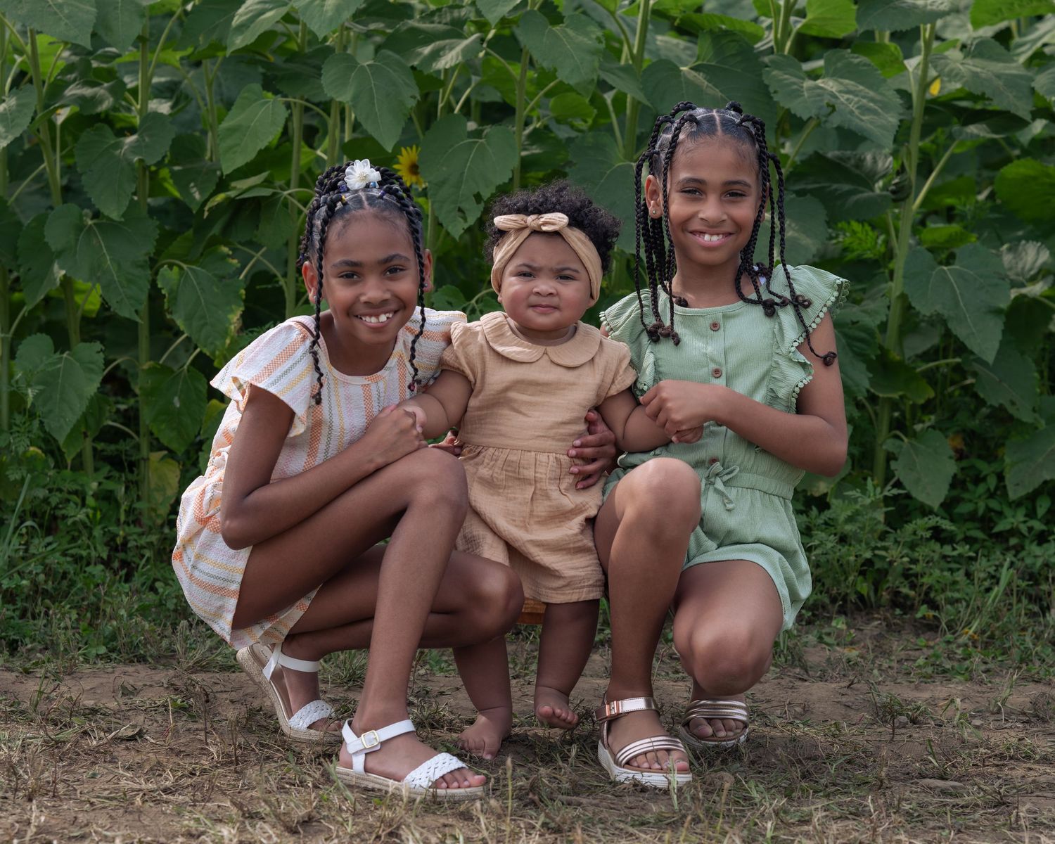 Three young sisters posing together in front of tall green sunflower plants in a garden setting.