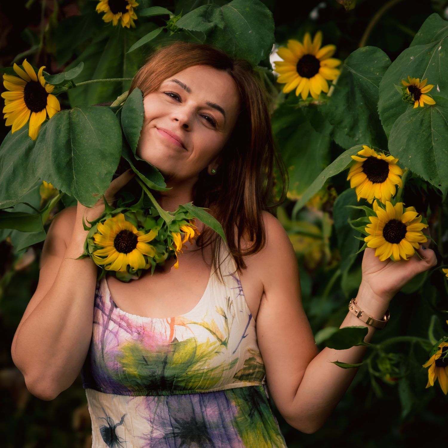 Woman smiling among sunflowers in a floral dress, surrounded by lush green leaves in a sunny garden.