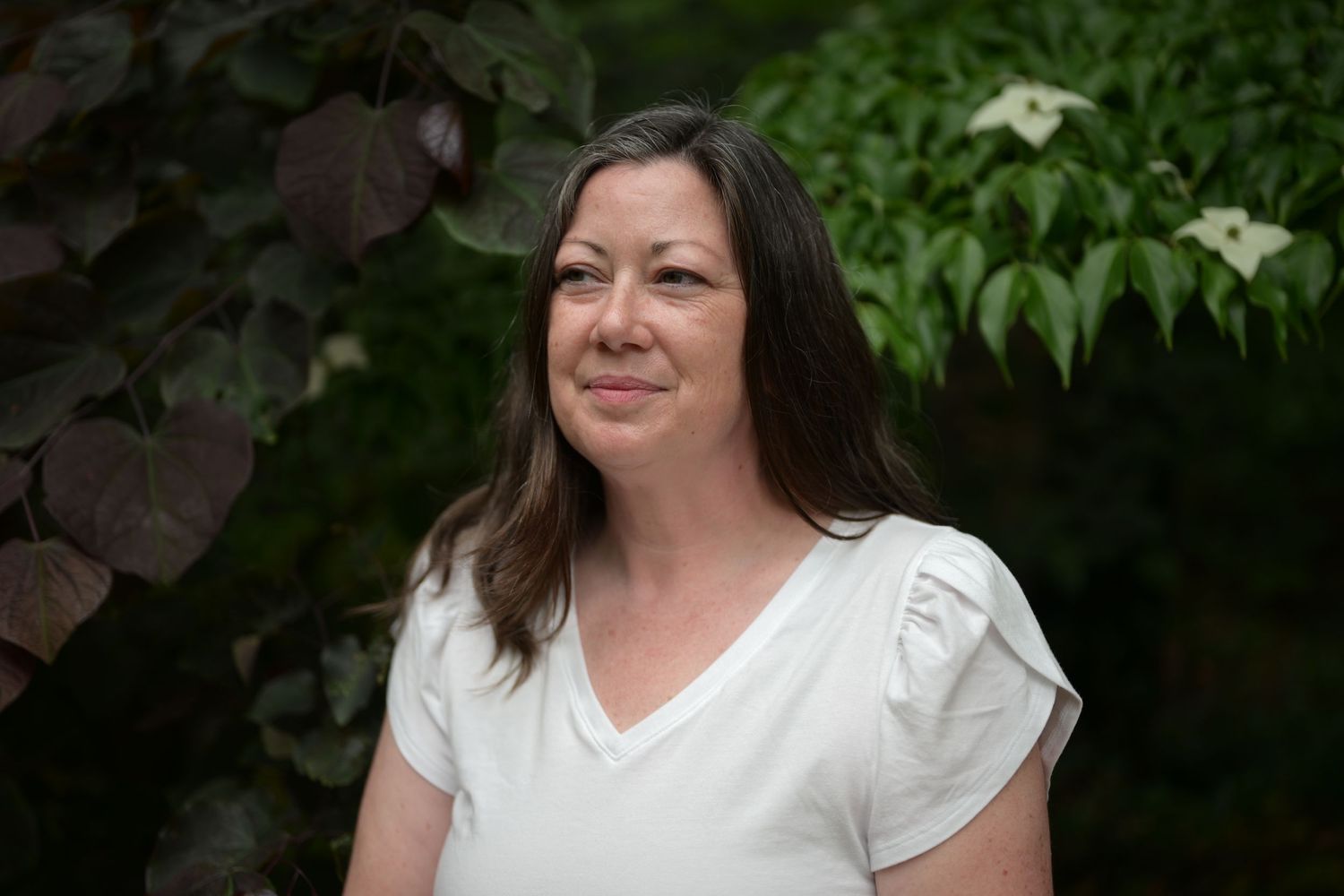 Woman in white top smiling outdoors surrounded by lush green and burgundy foliage.