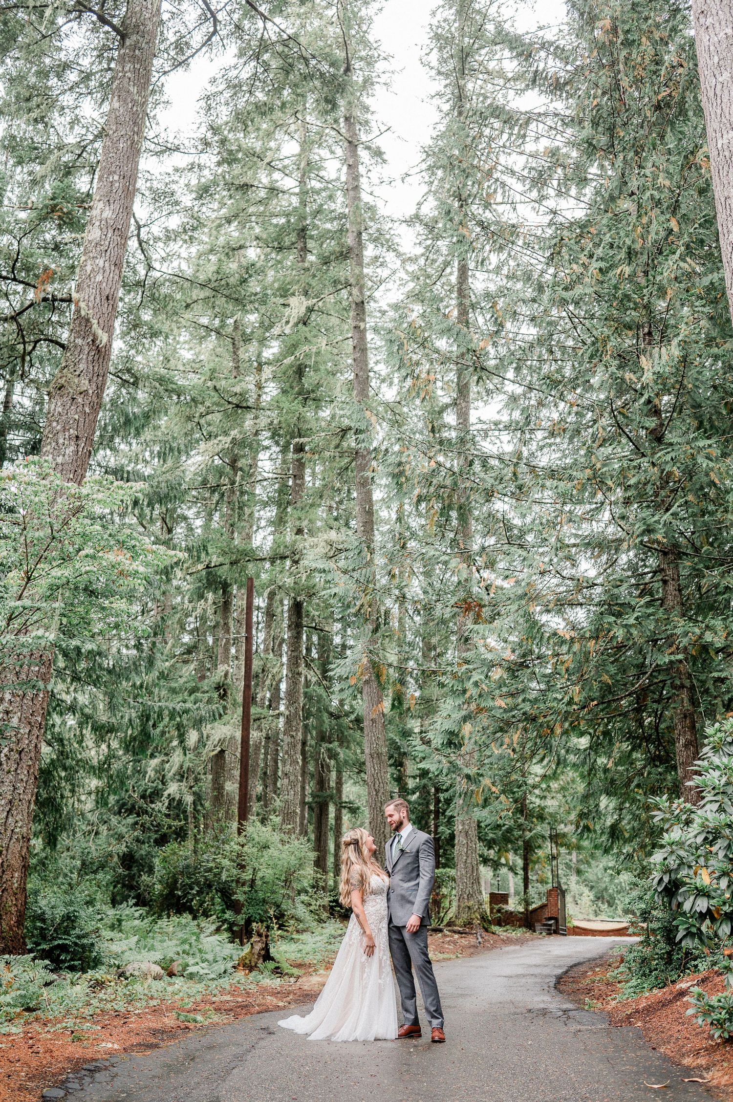 Bride and groom stand on a path surrounded by tall evergreen trees in a lush green woodland wedding setting. Photo by Pine + Vow