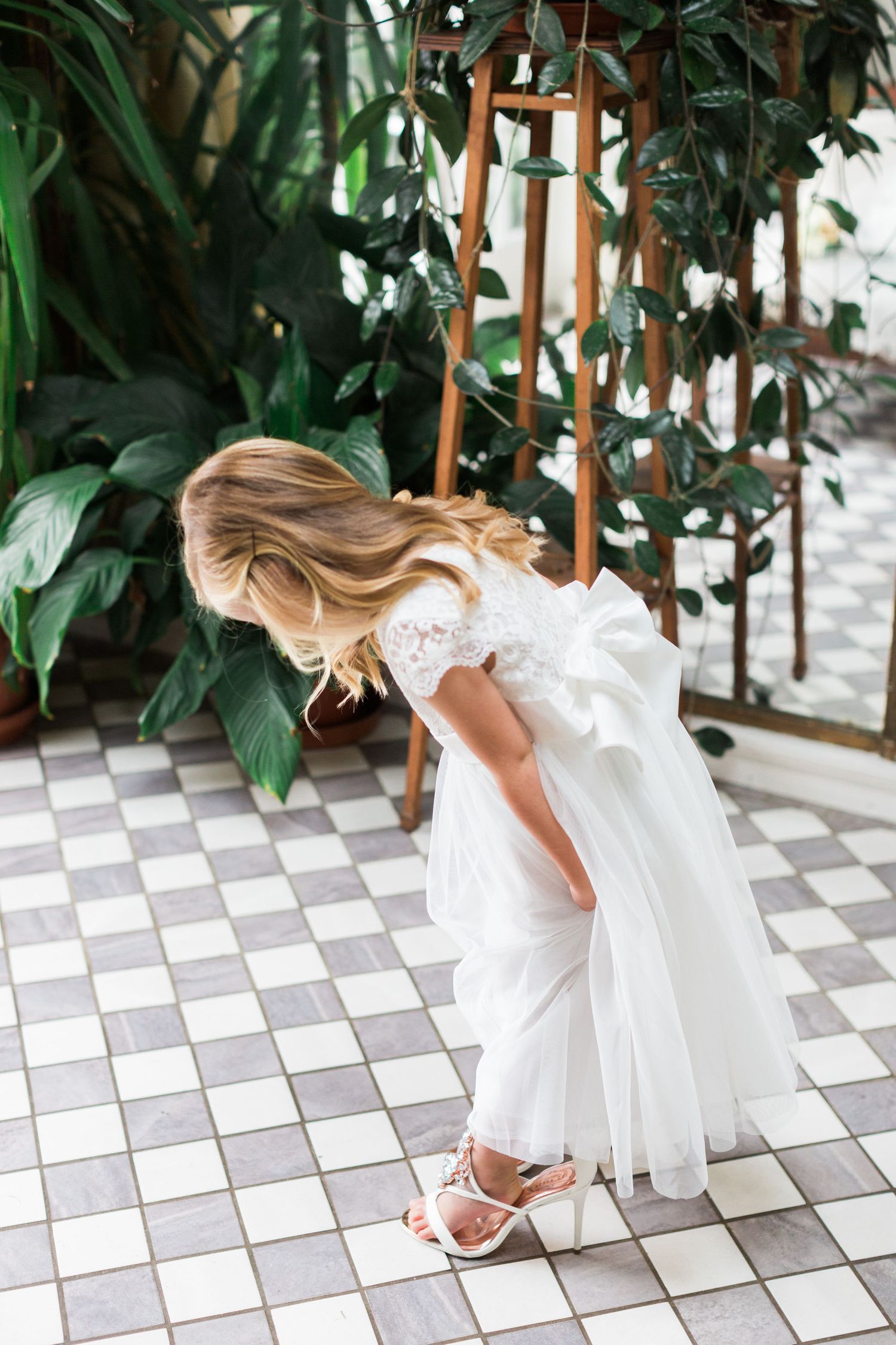 Young flower girl in white lace dress adjusting her sandal on checkered floor surrounded by lush green plants.