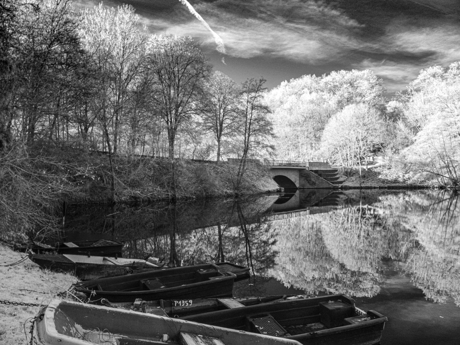 Black and white infrared photo of boats moored on a calm river near an arched stone bridge.