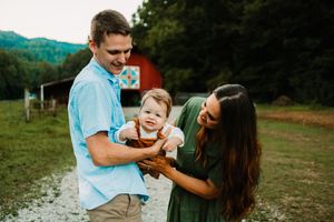 Young family with baby outdoors near red barn, parents smiling at infant in rustic countryside setting.